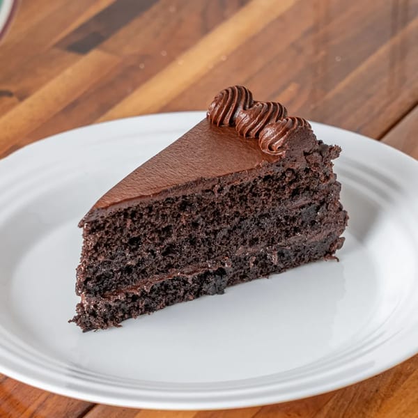 A slice of rich chocolate cake with chocolate frosting and decorative swirls on a white plate.
