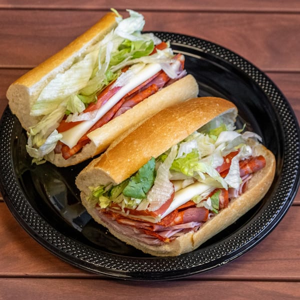 Two P&M Fine Foods deli subs, packed with meats, cheese, lettuce, and tomatoes on a black plate.