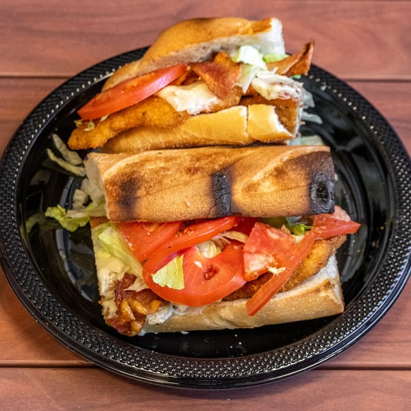 Two halves of a crispy chicken sandwich with bacon, lettuce, tomato, and sauce on a black plate.