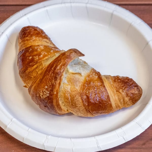 Golden-brown croissant with flaky layers and sugar crystals, served on a white paper plate.