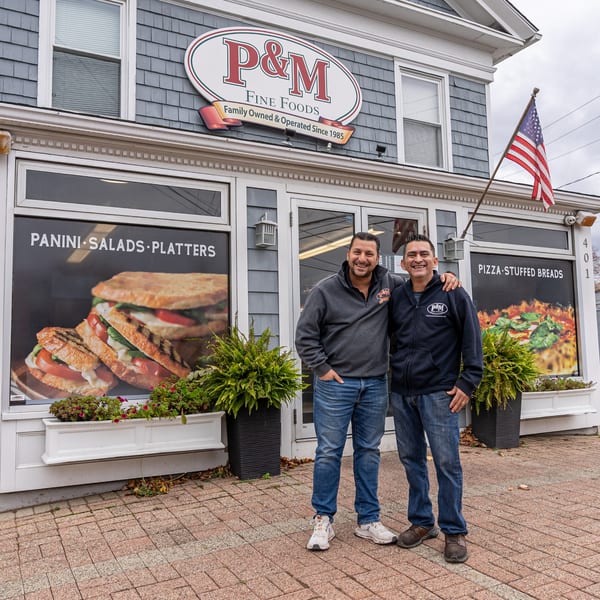 Two men smiling in front of P&M Fine Foods deli, with window displays of sandwiches and pizza.