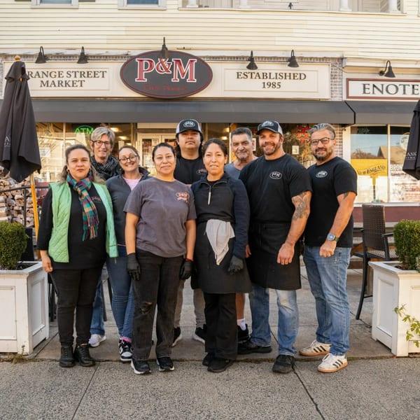 Nine P&M Fine Foods deli staff members stand smiling outside the restaurant entrance.