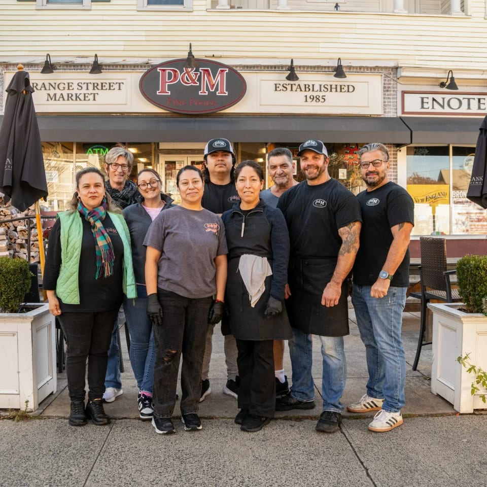 Nine P&M Fine Foods deli staff members stand smiling outside the restaurant entrance.