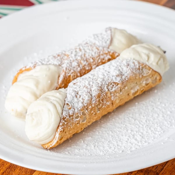 Two P&M Fine Foods cannolis, filled with cream and dusted with powdered sugar, on a white plate.