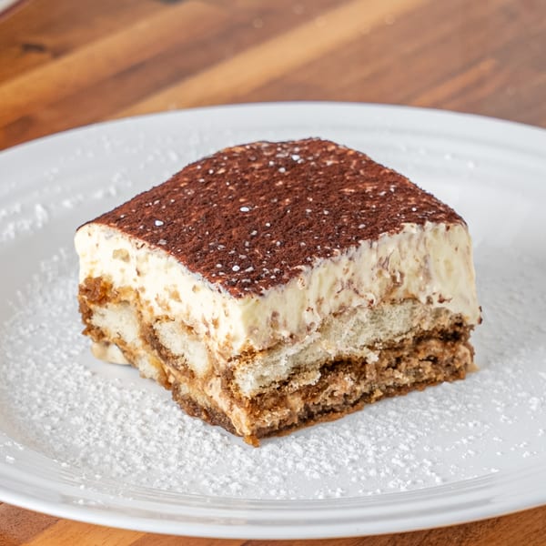 A slice of P&M Fine Foods tiramisu, dusted with cocoa powder and powdered sugar on a white plate.