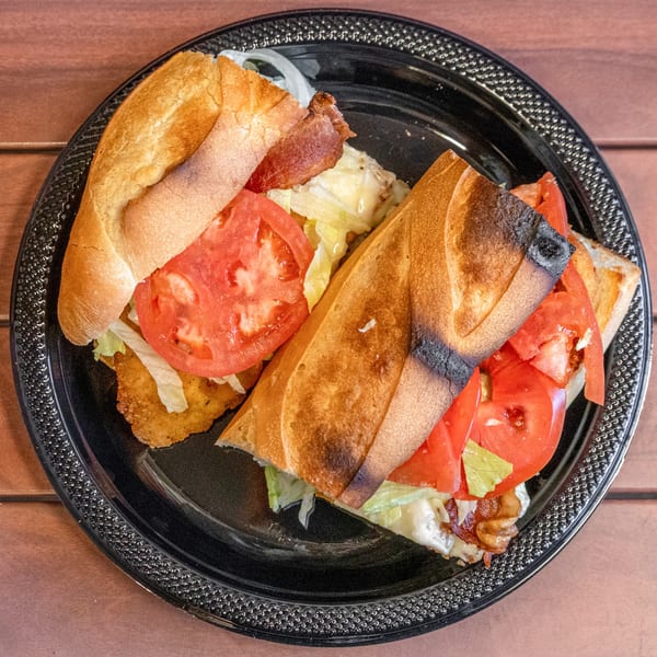Two halves of a deli sandwich with fried chicken, bacon, lettuce, and tomato on a black plate.