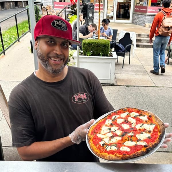 A smiling P&M Fine Foods employee in a red hat holds a freshly baked pizza with mozzarella and basil.